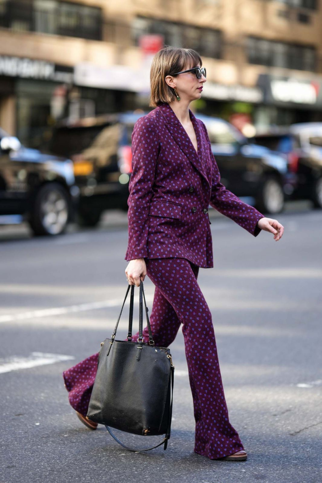 NEW YORK, NEW YORK - FEBRUARY 14: A guest wears black sunglasses, gold pendant earrings, a black / purple / burgundy print pattern blazer jacket, matching black / purple / burgundy print pattern flared pants, a black shiny leather handbag, dark brown shiny leather shoes , outside Brandon Maxwell, during New York Fashion Week, on February 14, 2023 in New York City. (Photo by Edward Berthelot/Getty Images)
