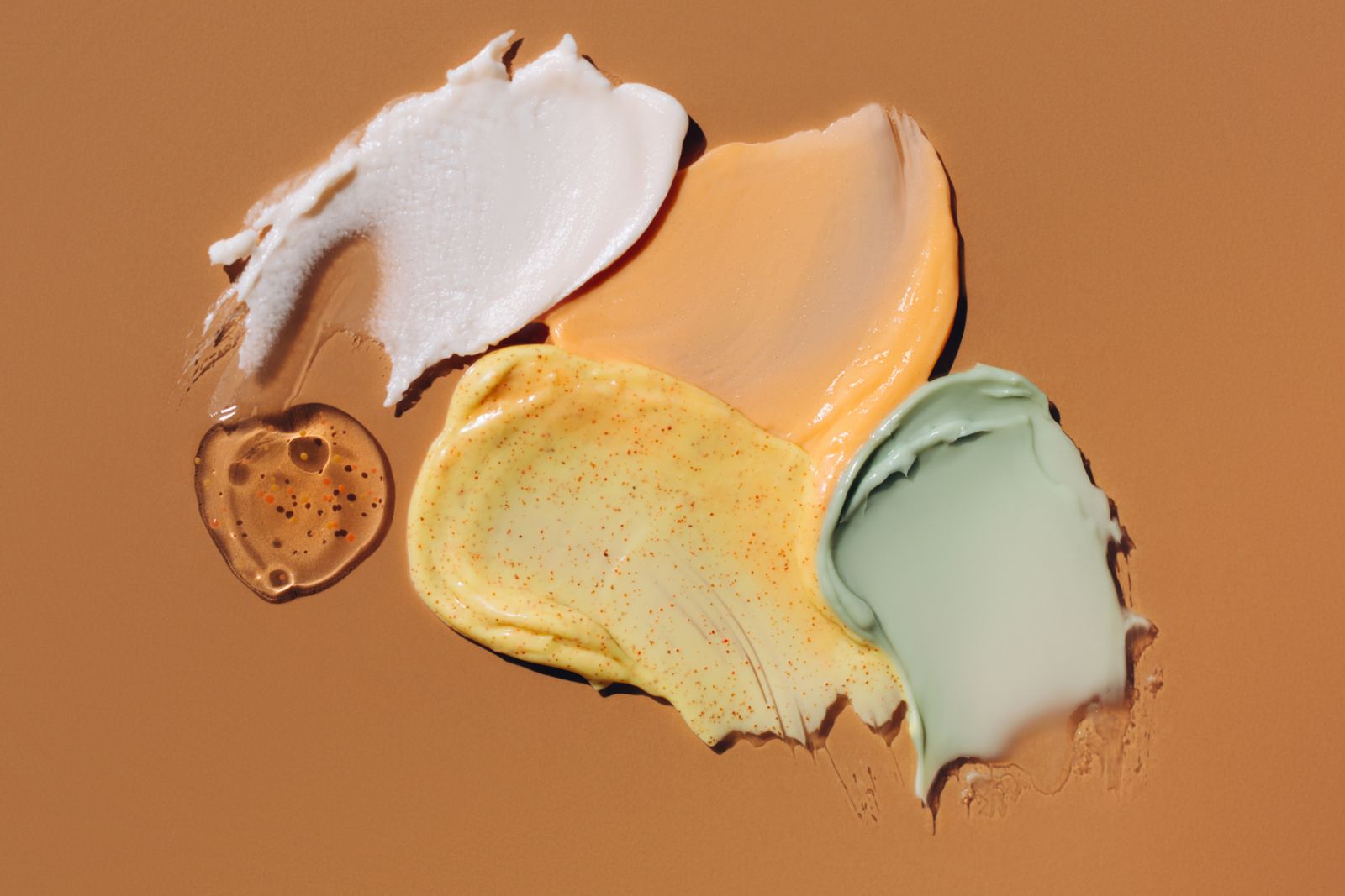 Set of multicolored cosmetic smears. White, orange and green creams, yellow scrub with exfoliating particles and transparent blob of moisturising gel with particles on brown background. Concept of trendy body care procedures of the year