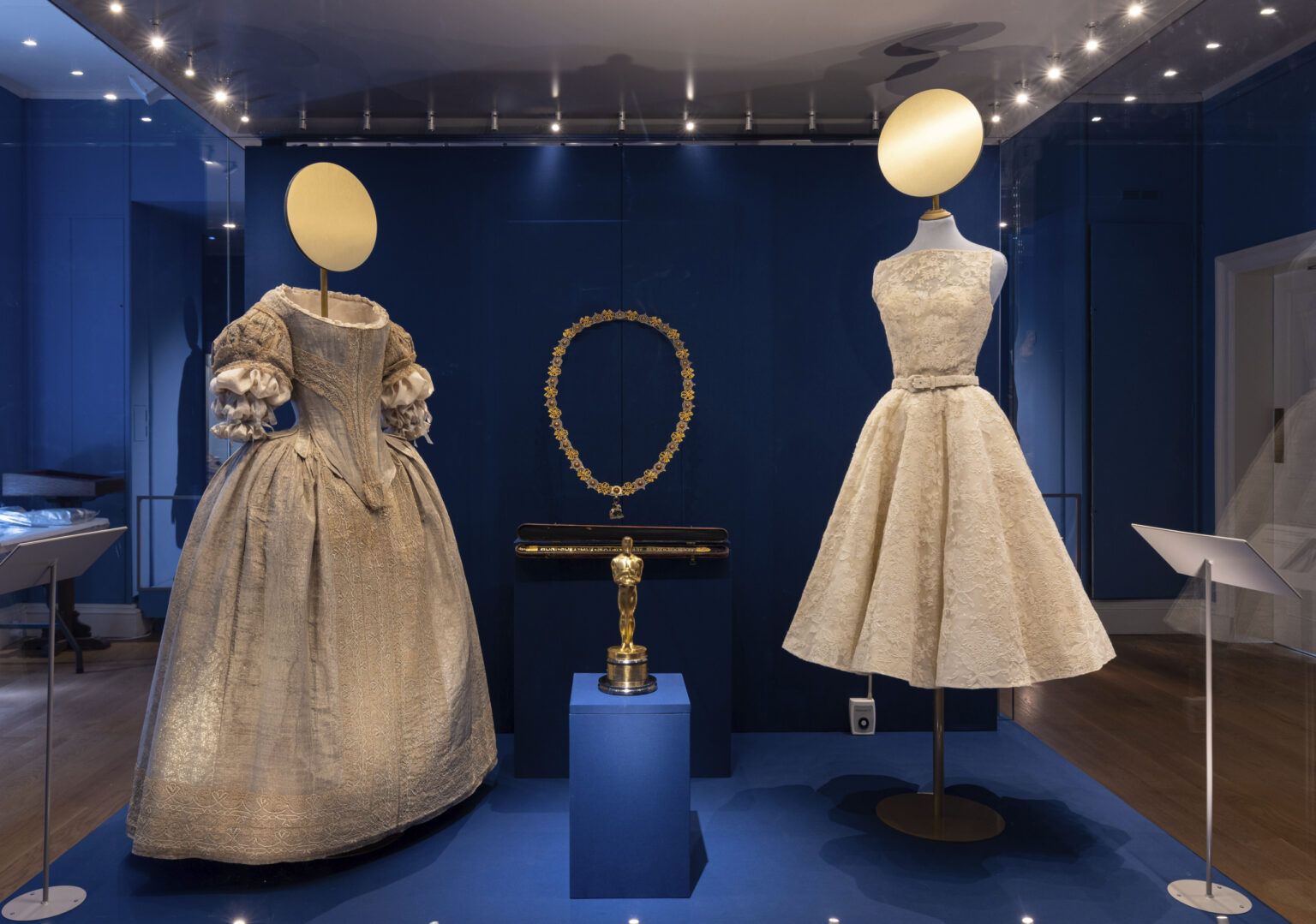 The Pigott Gallery (Room 2), displaying a 1660s silver tissue gown worn by Lady Theophilia Harris (d.1702) next to a dress designed by Edith Head for the film Roman Holiday, altered by Givenchy for the Oscars 1954 and worn by Audrey Hepburn. Also displayed is the Oscar Statuette presented to Edith Head at the 1954 Academy Awards for Costume Design for Roman Holiday.Crown to Couture, a major exhibition at Kensington Palace, examines and celebrates how fashion from the royal Georgian court has inspired red-