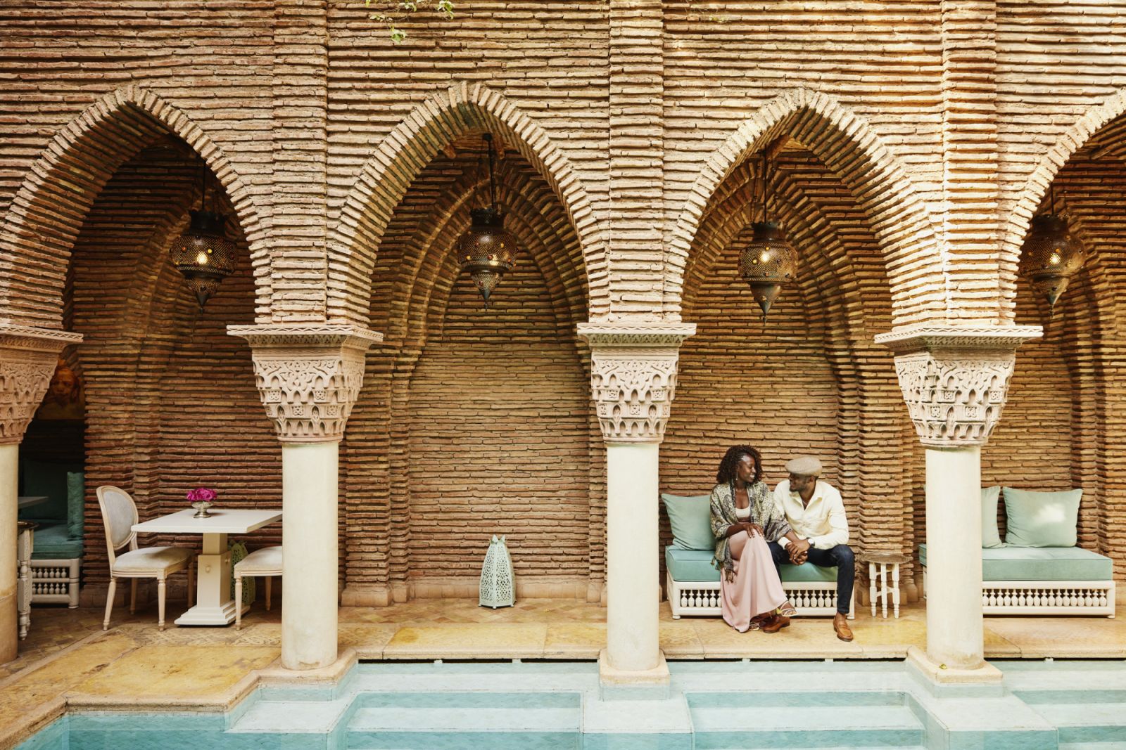 Wide shot of smiling couple holding hands while sitting by pool in courtyard of luxury hotel while on vacation