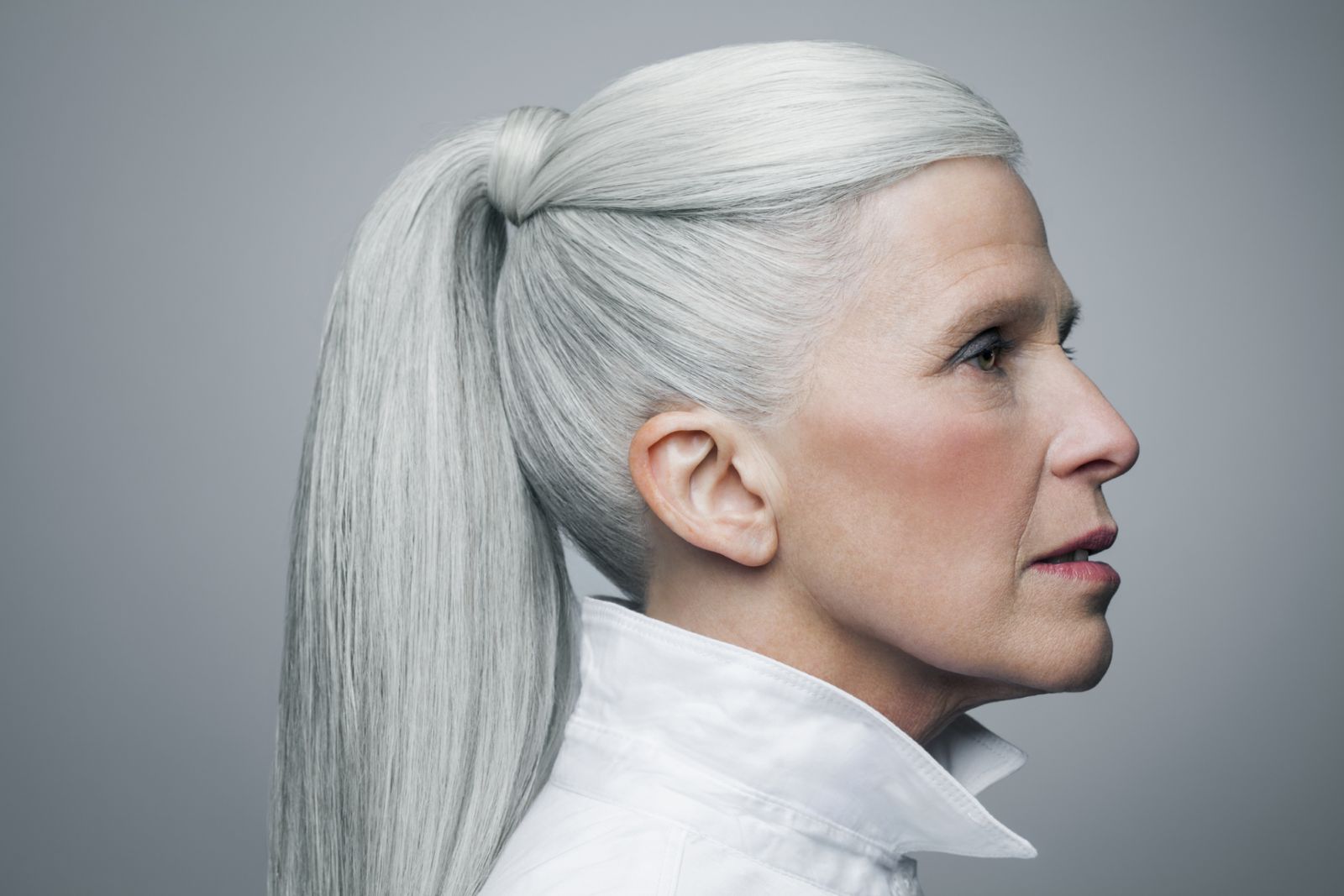 Mature woman with long, straight, silvery, grey hair in a ponytail wearing a white shirt in front of a grey background in a profile position, portrait.