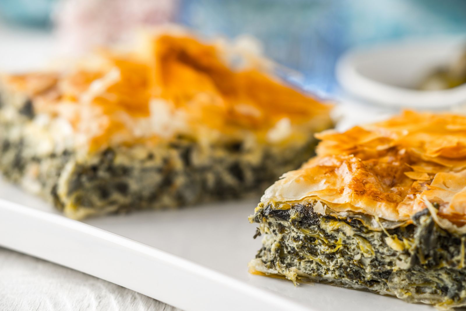 Greek pie spanakopita on the white plate on the white wooden table with blurred accessorizes horizontal