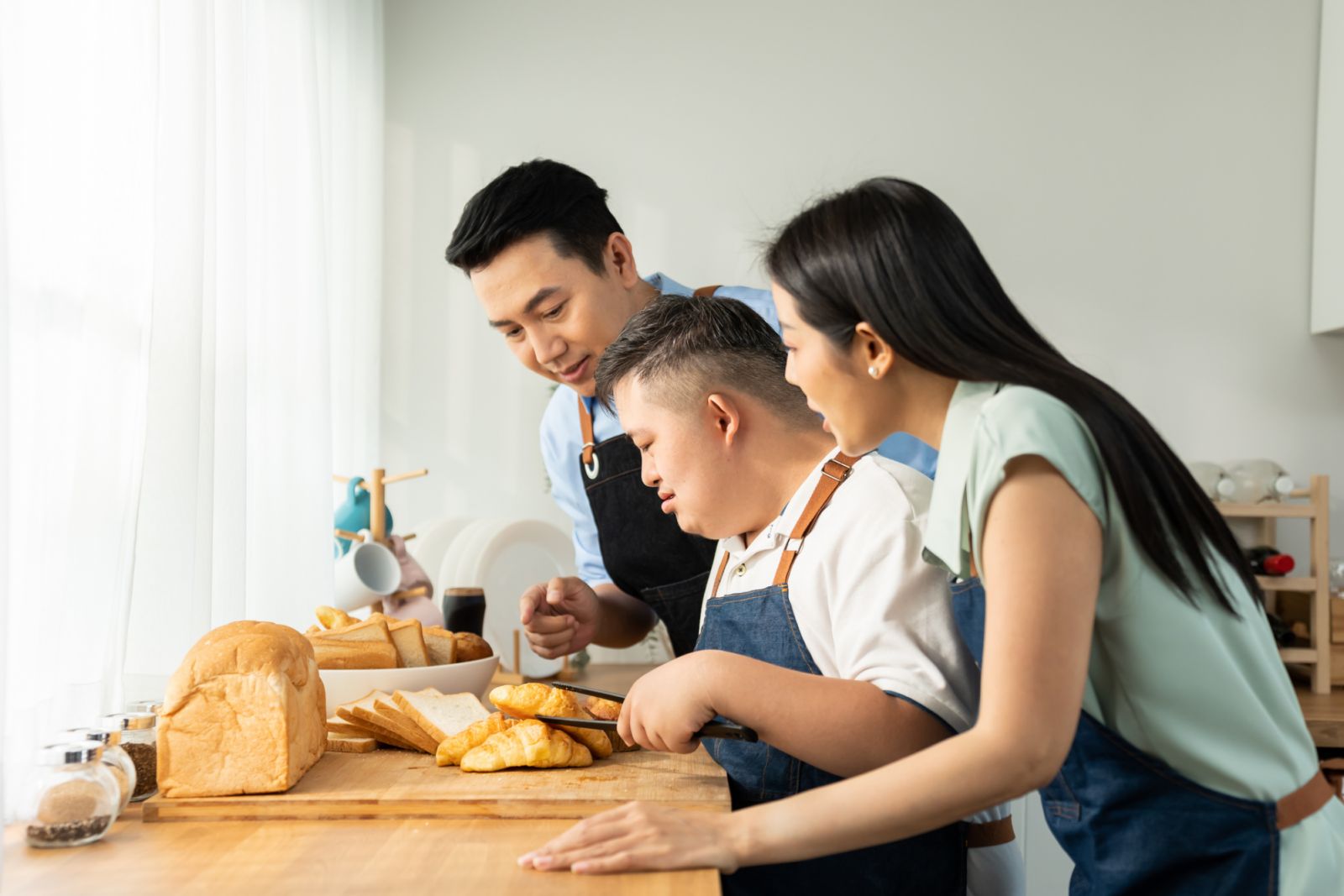 Asian happy family baking bakery with down syndrome's son in kitchen. Beautiful loving mother and father take care and teaching young man with autism or special needs cooking foods indoors at home.