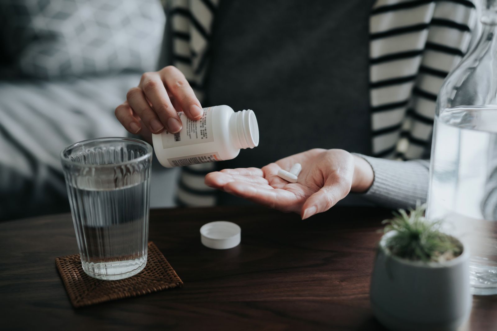 Close up of young Asian woman holding a pill bottle, pouring pills into palm of hand, with a glass of water on the side. Medicine, healthcare and people concept