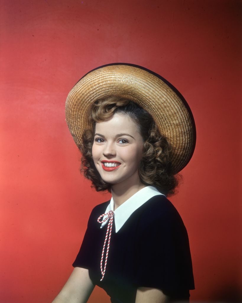circa 1945: Smiling studio portrait of American actor Shirley Temple, sitting in front of a red backdrop, wearing a straw hat and black top with white collar. (Photo by Hulton Archive/Getty Images)