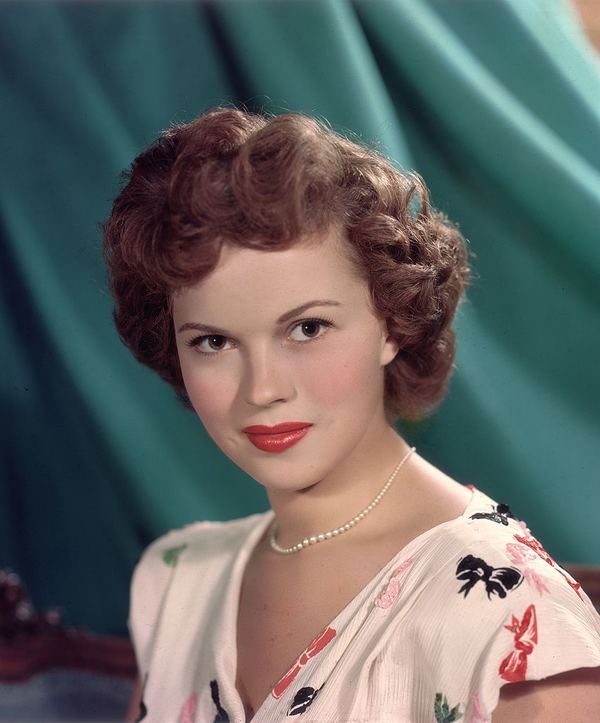 circa 1955: Headshot studio portrait of American actor Shirley Temple, in front of a green backdrop, wearing a short sleeve white blouse with a colored bows pattern and a pearl necklace. (Photo by Hulton Archive/Getty Images)
