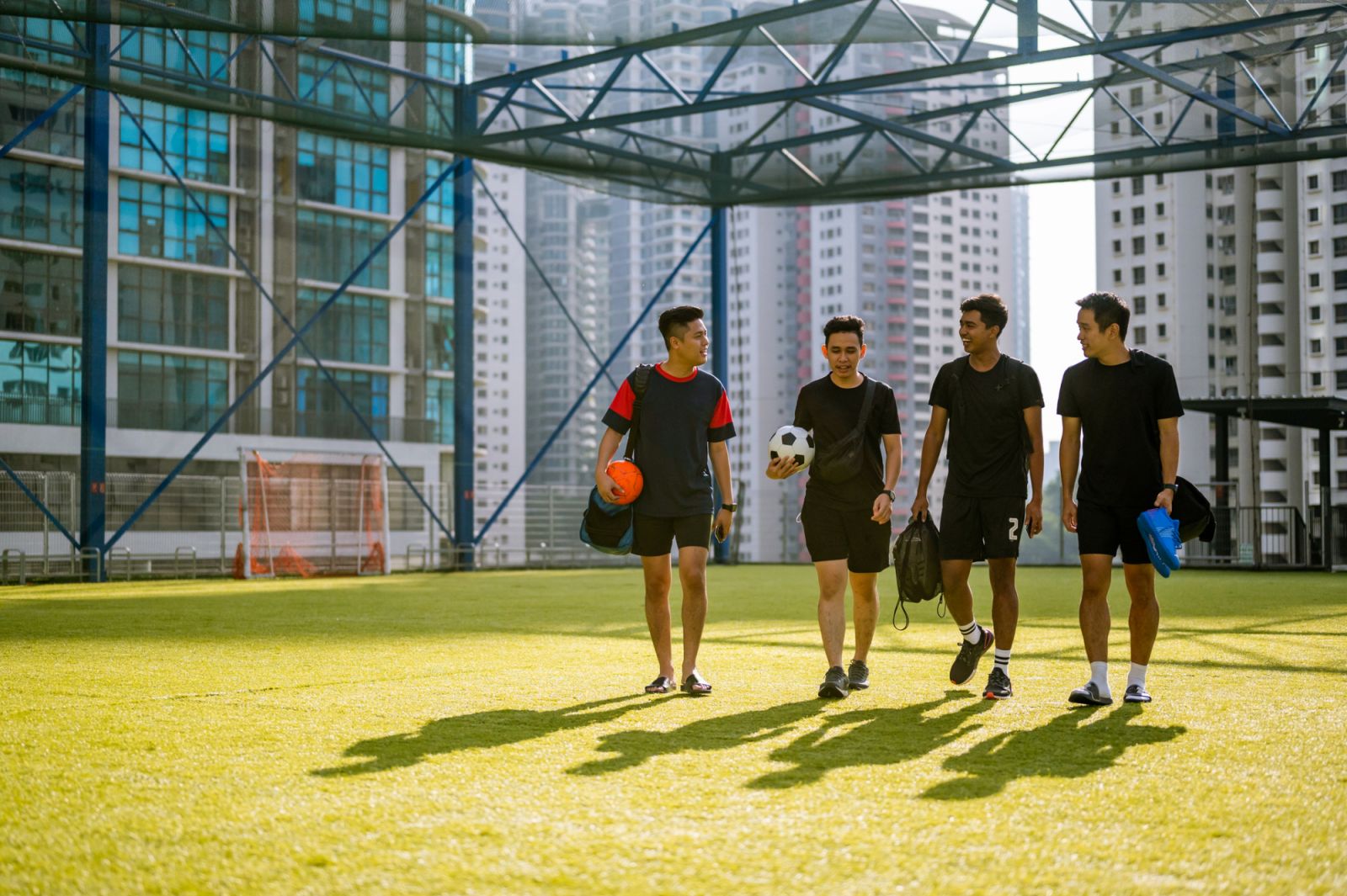 Happy young Asian male football team walking onto sports field with gear for morning practice.