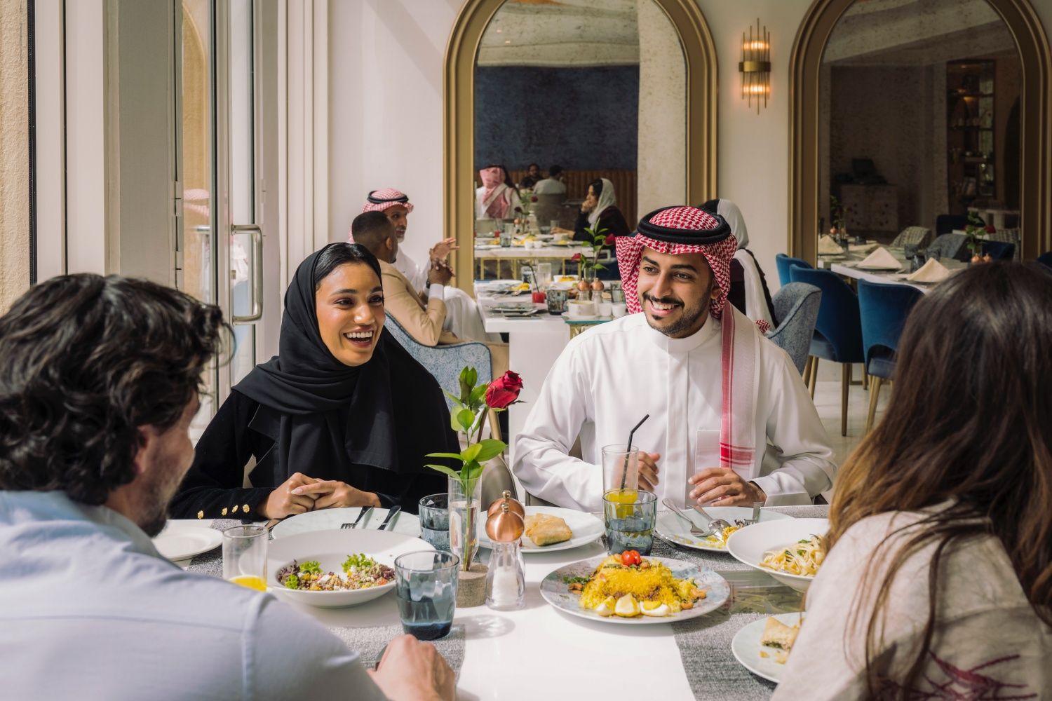 Waist-up view of smiling men and women in traditional and western attire sitting face to face at window table and conversing over midday meal.