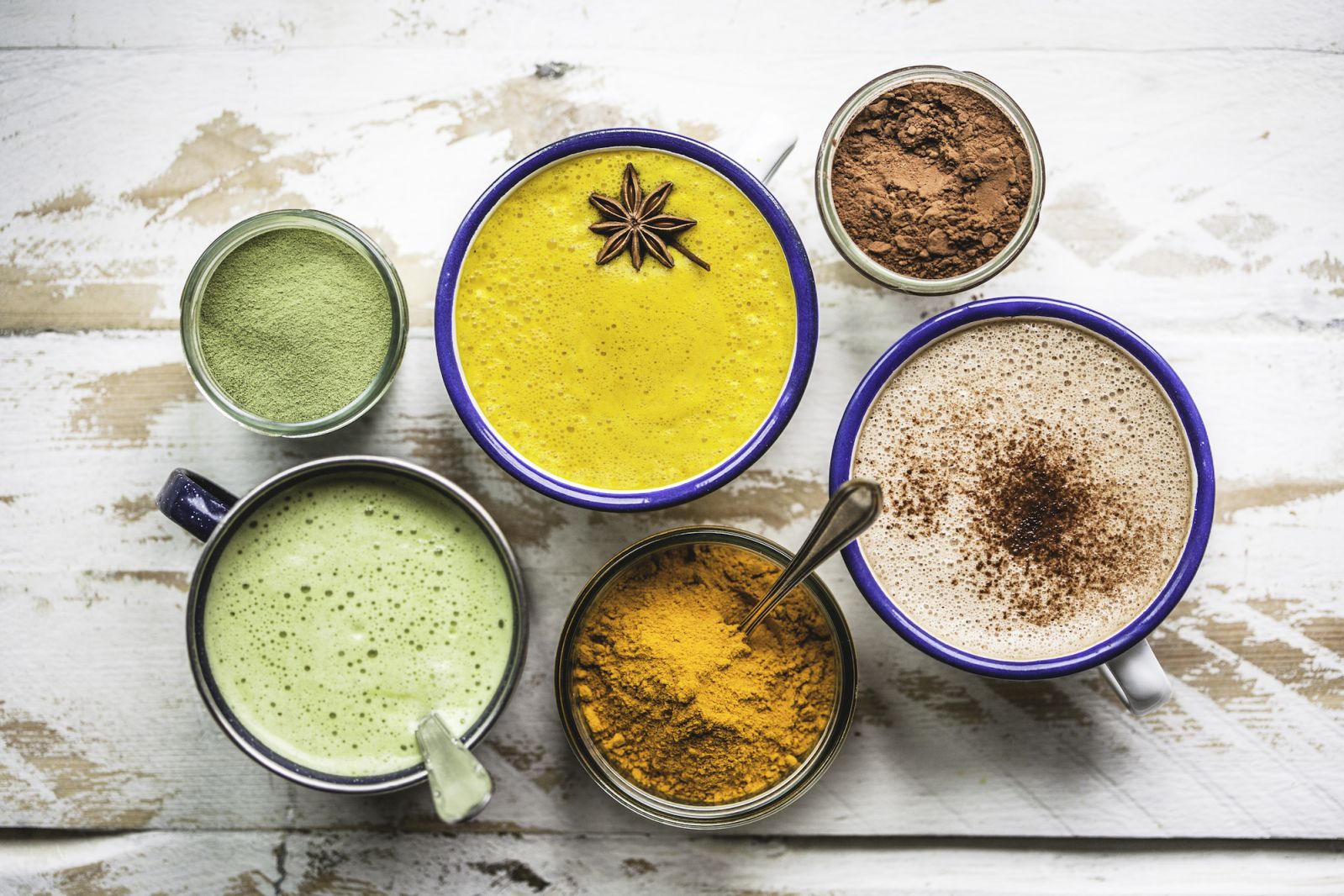 A Matcha Latte, a Golden Milk Latte and a Raw Cacao Hot Chocolate assembled in a cluster on a rustic white wooden table top, with jars of the ingredients gathered around.