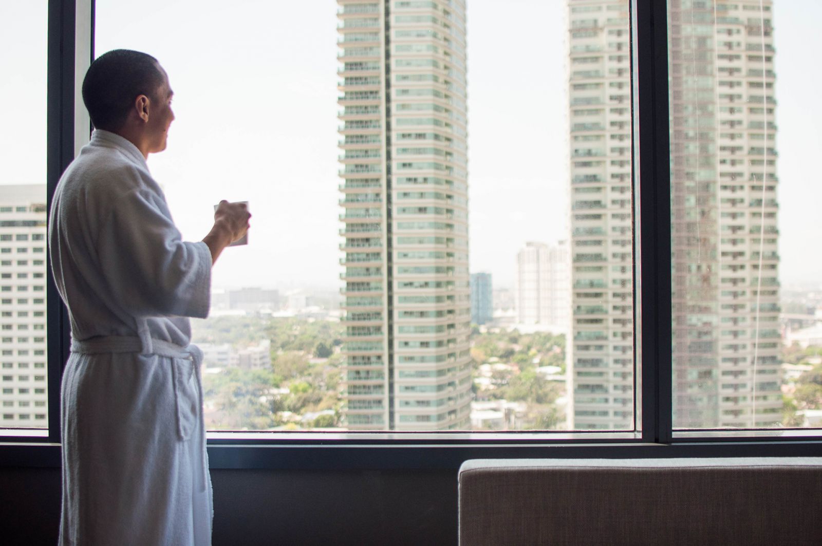 A young man is wearing a bathrobe while drinking a cup of hot coffee over a large window overseeing the city