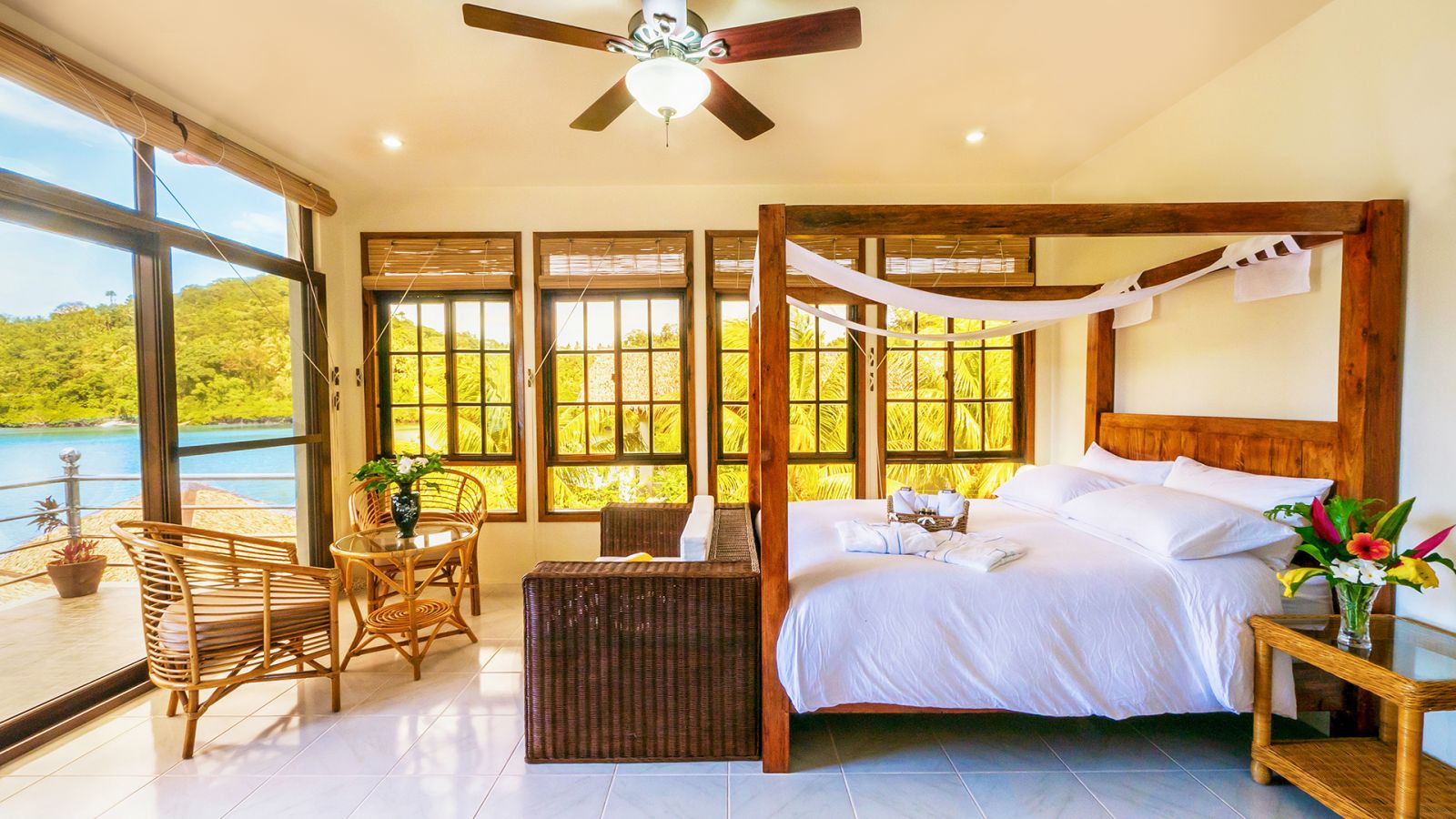 Wooden four-poster bed with white bedding, white floor tiles, rattan tables, chairs, and sofa. Ceiling fan. Floor to ceiling windows and sliding doors leading to waterfront balcony. Fresh, clean and calming room atmosphere.