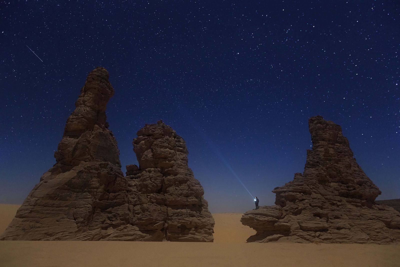 A man standing on a rock feature in the desert, shining his torch into the stary night sky