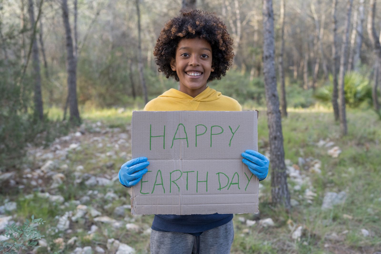 A sweet African American boy holds up a "Happy Earth Day" sign as he proudly cleans up the forest around his. He is dressed casually and wearing gardening gloves as she smiles happily.