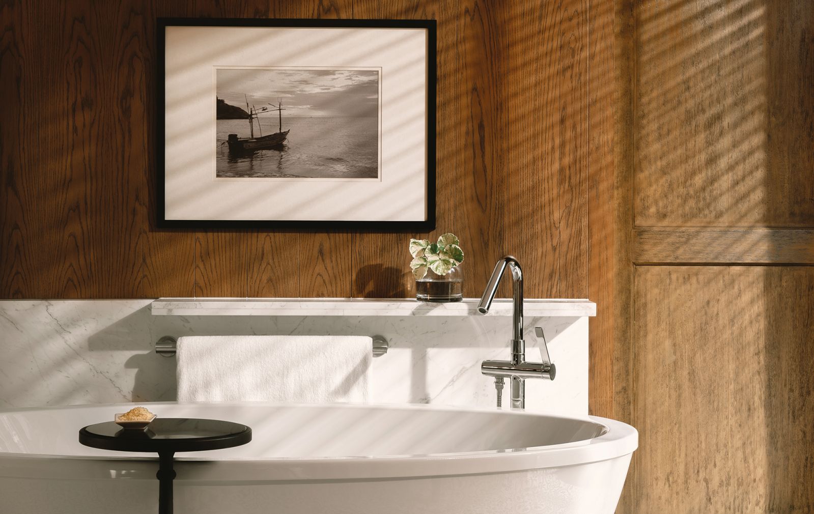 The guest bathroom at Kimpton Kitalay on Koh Samui, Thailand features a freestanding bathtub and a curated floor-mounted faucet in chrome in front of a natural wood wall to give the hotel bathroom a feeling of warmth and back to nature aesthetic