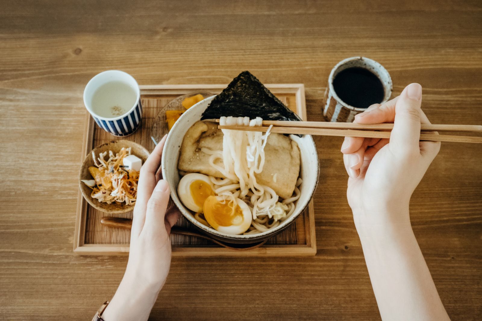 Point of view shot of woman's hand enjoying freshly made traditional Japanese udon soup noodles with salad and tea in a restaurant