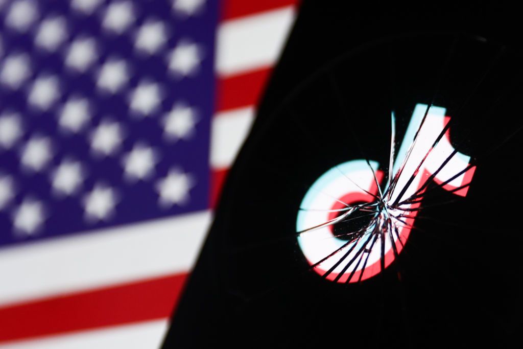 TikTok logo displayed on a phone screen is seen through the broken glass with American flag displayed on a screen in the background (Photo: Jakub Porzycki/NurPhoto via Getty Images)
