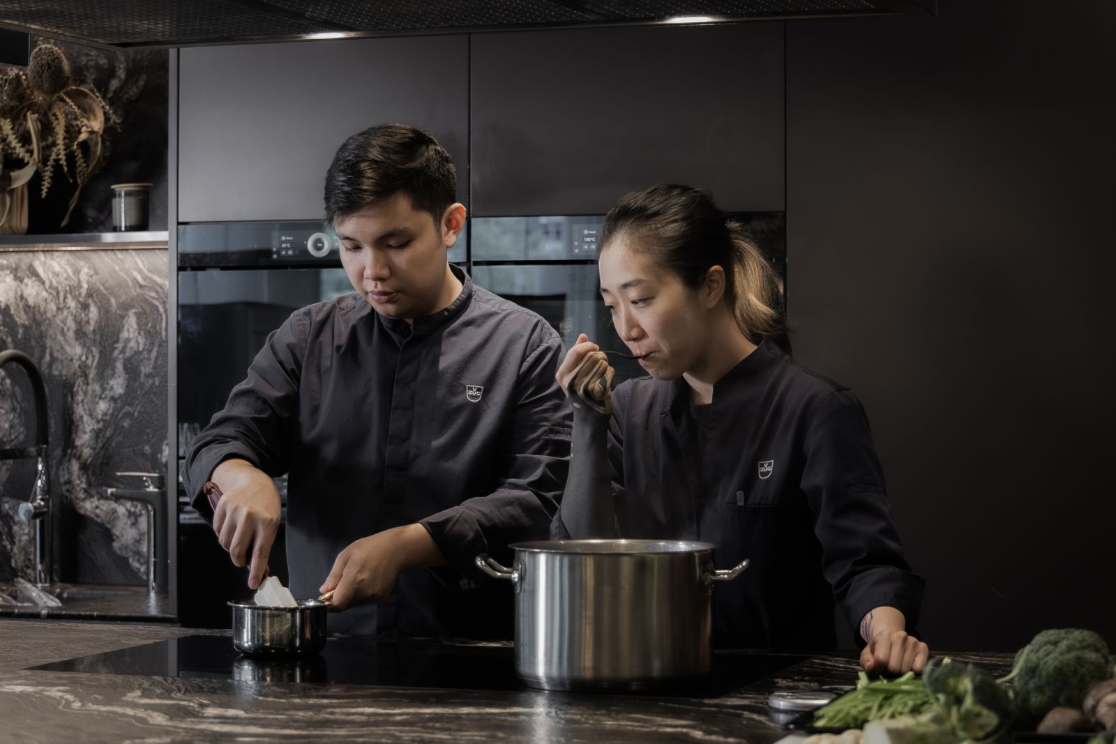 Chef Ng doing a taste test while Chef Ow tends to the dish on stove