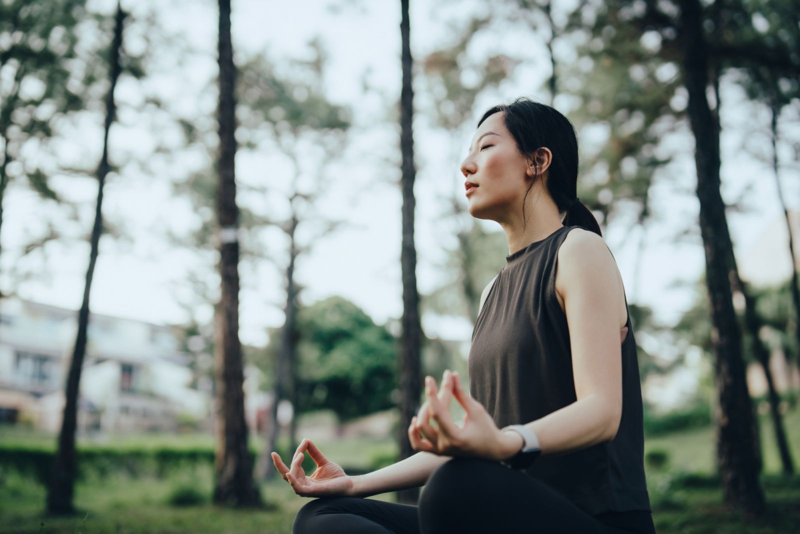 Relaxed young Asian woman with eyes closed sitting on exercise mat outdoors in park, doing yoga meditation exercise in lotus position. Fitness and wellness. Exercise outdoors. Healthy living lifestyle