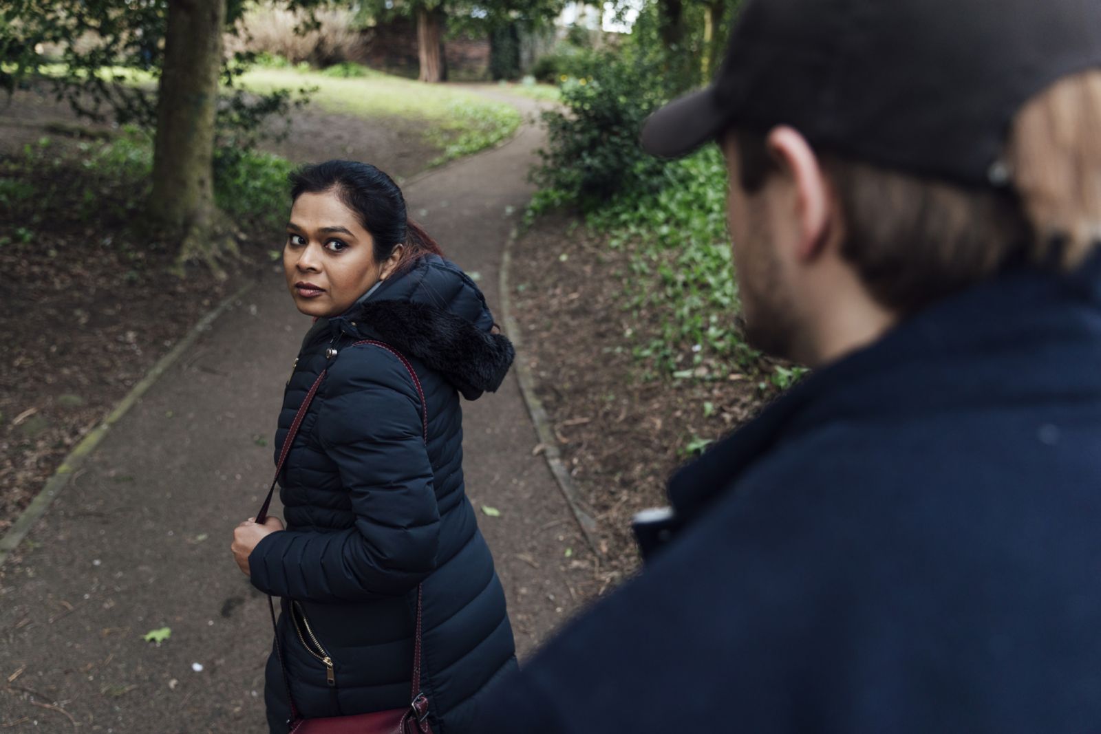 An over-the-shoulder view of a woman who looks terrified as she walks through a public park in Hexham in the North East of England. She is looking over her shoulder and can see she is being followed by a man who is hiding part of his face under a cap leaving her vulnerable and scared.