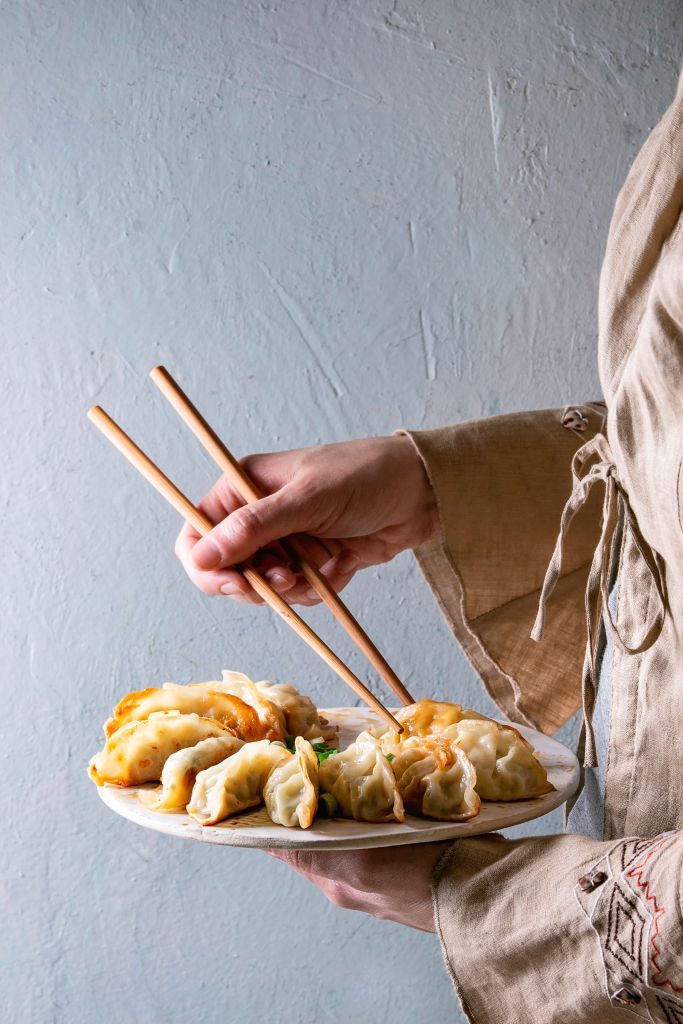 Fried asian dumplings Gyozas potstickers on white ceramic tray and wooden chopsticks in female hands, Grey wall at background, Asian dinner. (Photo by: Natasha Breen/REDA&CO/Universal Images Group via Getty Images)