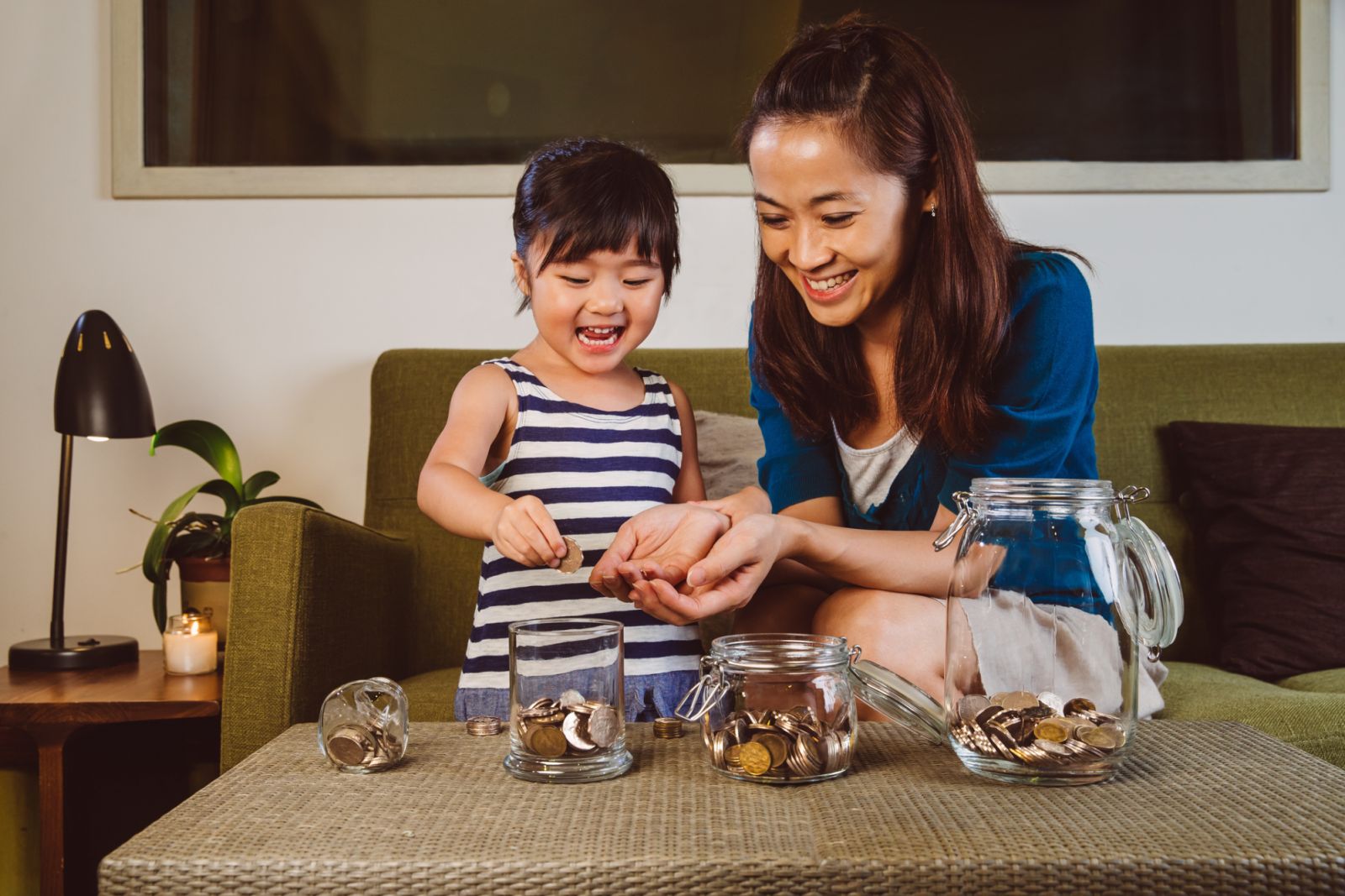 Lovely little girl putting coins into different glass jars on the coffee table joyfully under her pretty young mom's guidance at home.
