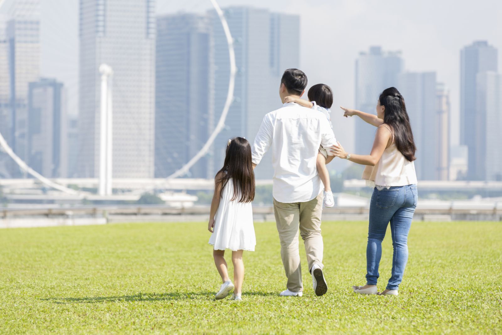 Mom and dad with their two children walk together in a city park. A city skyline and ferris wheel are in the background.