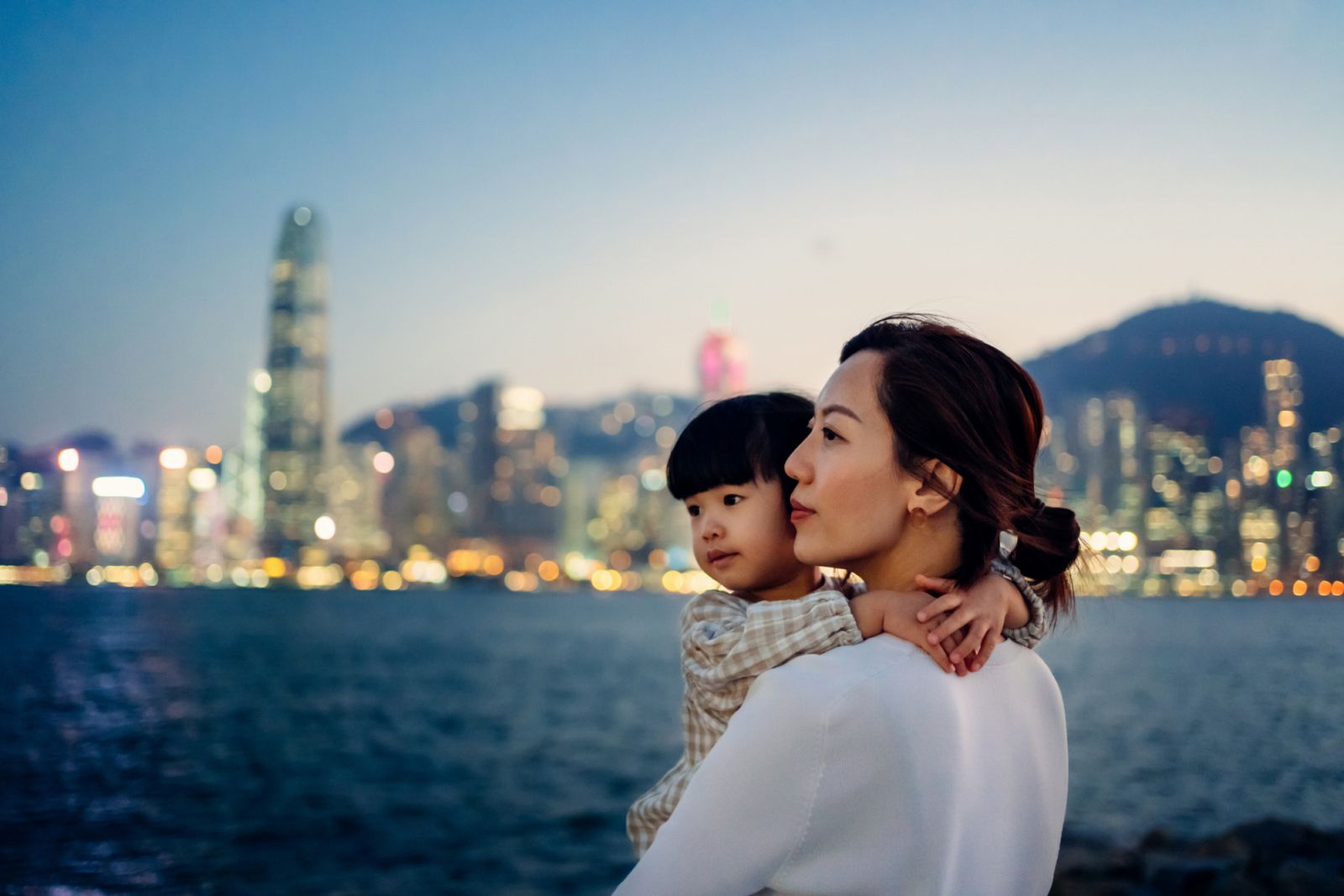 Loving young Asian mother carrying adorable little daughter in her arms, looking away while standing against the promenade of Victoria harbour and illuminated urban city skyline. Enjoying intimate mother and daughter bonding time together