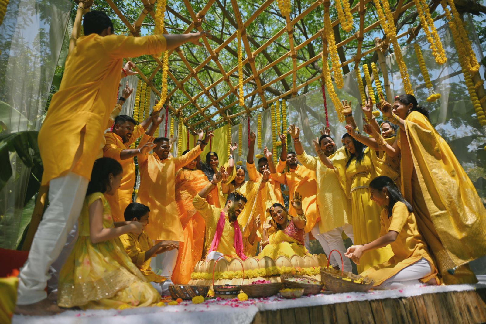 Family and friends dressed beautifully in a yellow attire gather around the bride and groom during their haldi ceremony, dancing joyously to the lively beats. With laughter echoing and hands clapping, they create a circle of love, warmth and support. Each step is a testament to the shared happiness, marking the beginning of a new journey filled with love, laughter, and cherished memories