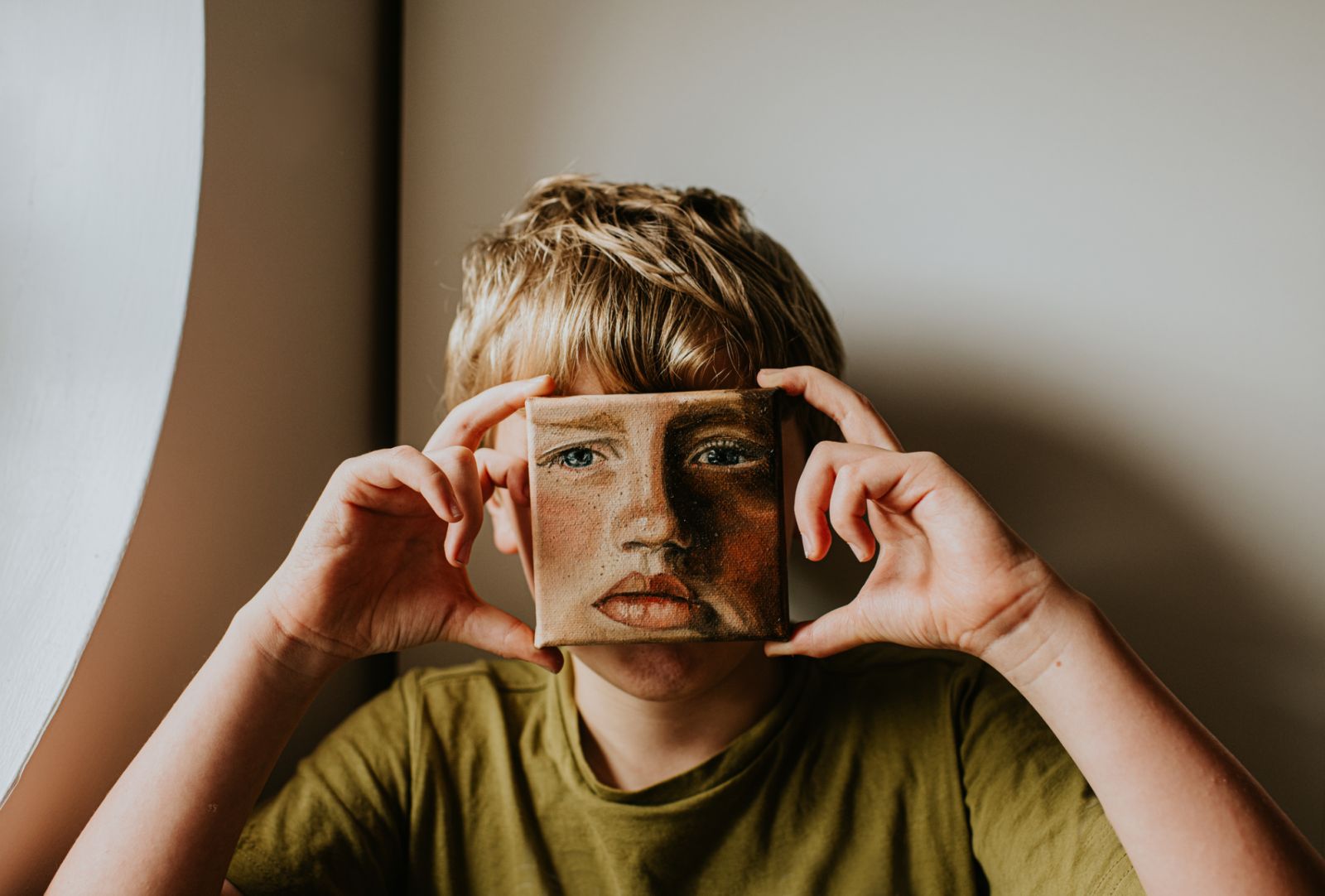 A young lad covers his face with a small oil painting of himself. The expression on the painting is serious. Conceptual with space for copy.