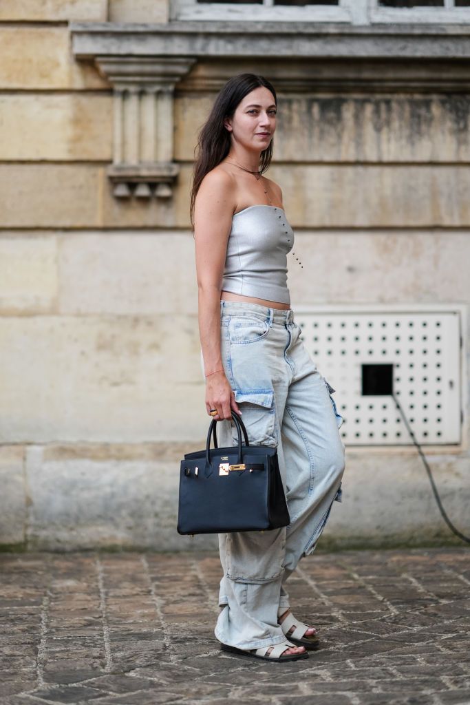 PARIS, FRANCE - JUNE 27: Alice Mouzon wears black beaded silver necklace, silver metallic bandeau top, washed light blue low-rise denim jean cargo pants, white Hermes chypre sandals leather shoes, black Hermes Birkin leather bag, gold rings, outside Ashi Studio, during the Paris Haute Couture Week Fall/Winter 2024-2025 on June 27, 2024 in Paris, France. (Photo by Edward Berthelot/Getty Images)