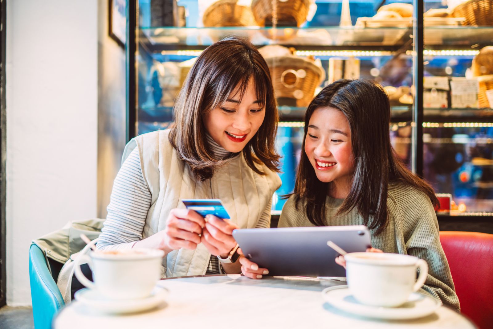 A mother shares the world of e-banking with her Gen-Z daughter, guiding her through financial apps on a tablet. Their cozy cafe setting is filled with warmth and shared learning, as they explore the realms of digital finance together over cups of coffee and the inviting display of a bakery.