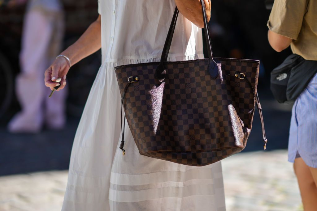 COPENHAGEN, DENMARK - AUGUST 11: A guest wears a silver bracelet, a white latte long dress, a dark brown and brown checkered print pattern in coated canvas Neverfull handbag from Louis Vuitton , outside Mark Kenly Domino Tan during Copenhagen Fashion Week Spring/Summer 2023, on August 11, 2022 in Copenhagen, Denmark. (Photo by Edward Berthelot/Getty Images)