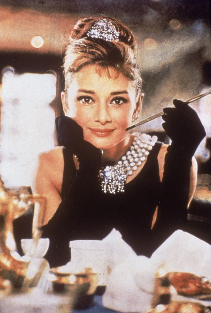 Belgian-born actress Audrey Hepburn (1929 - 1993), in a black, shoulderless dress, matching gloves, and a tiara, smiles with a cigarette holder in her hand, in her role as Holly Golightly the film, 'Breakfast at Tiffany's,' directed by Blake Edwards, New York, New York, 1961. (Photo by Paramount Pictures/Courtesy of Getty Images)