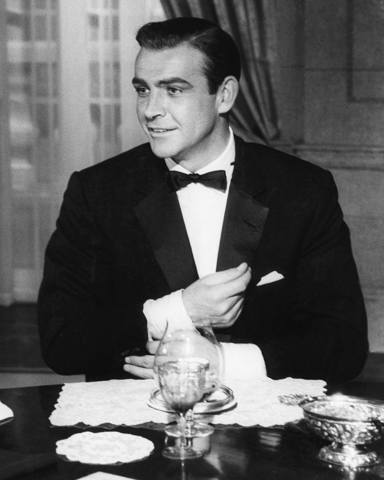Sean Connery, in a scene from Goldfinger, adjusts a cuff of his tuxedo while dining with M and the Prime Minister.