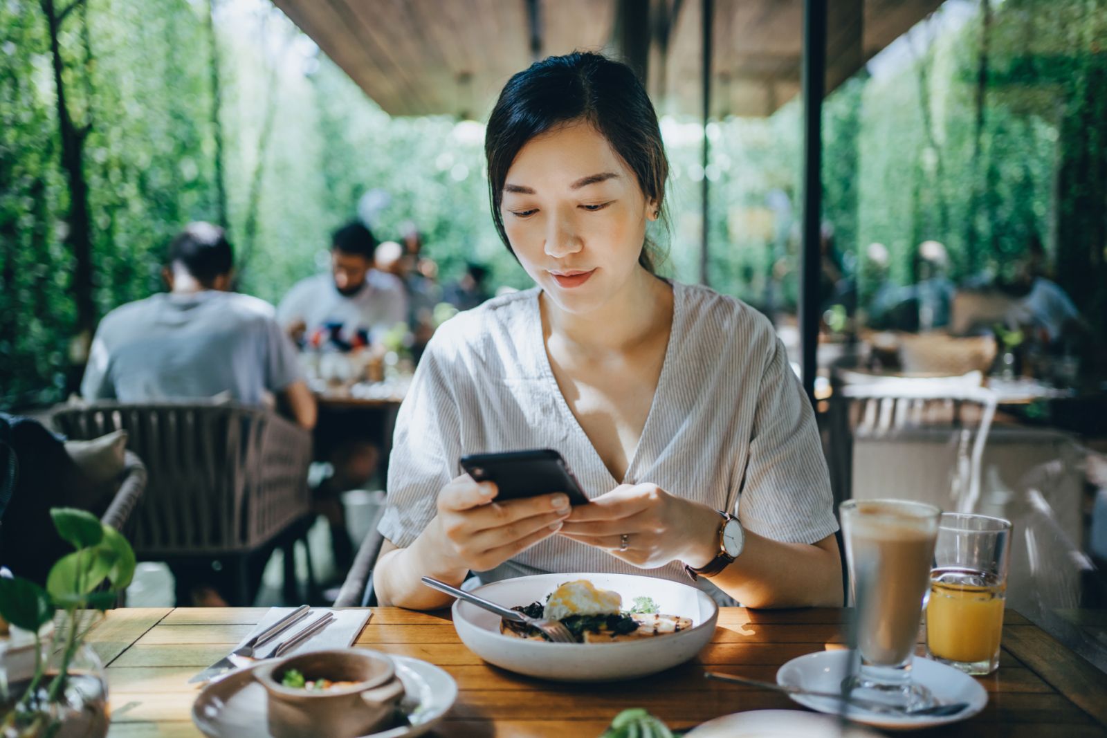 Smiling young Asian woman using smartphone while having brunch in an outdoor restaurant surrounded by lush foliage and beautiful sunlight. Lifestyle and technology