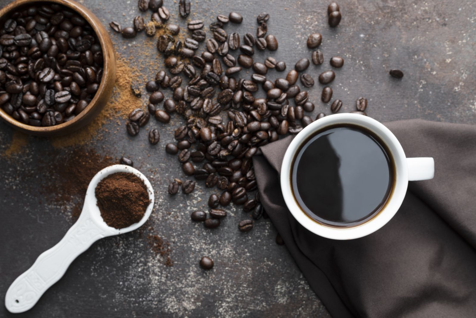 Direct above view of roasted coffee beans, white coffee cup with black coffee, dark brown colored napkin with one white spoon full with blended coffee on dark brown background