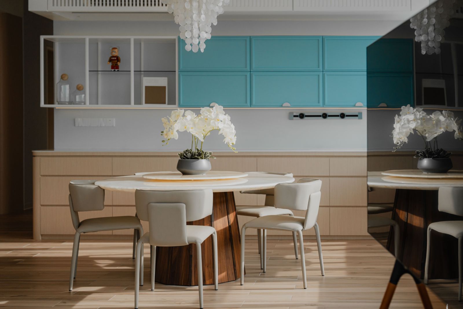Modern dining area featuring a round white marble table with wooden base, light grey upholstered chairs, and turquoise upper cabinets.