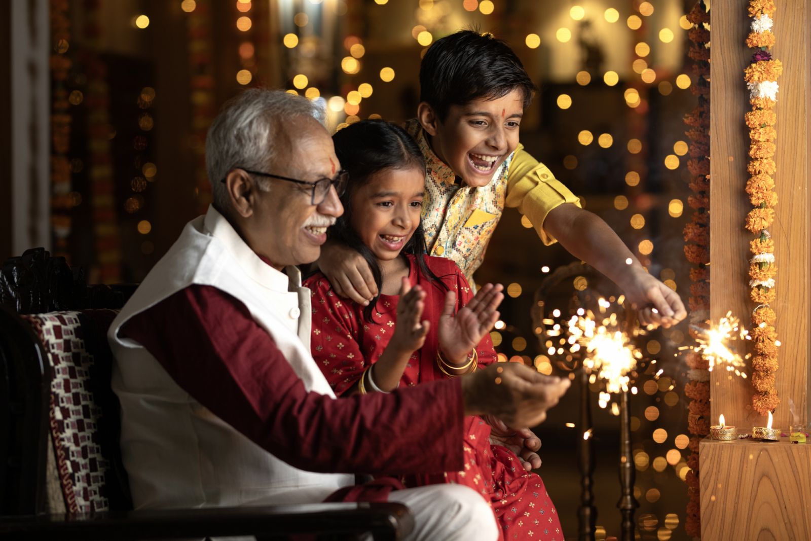 Cheerful grandfather and grandchildren playing with sparklers while celebrating Diwali festival at illuminated home