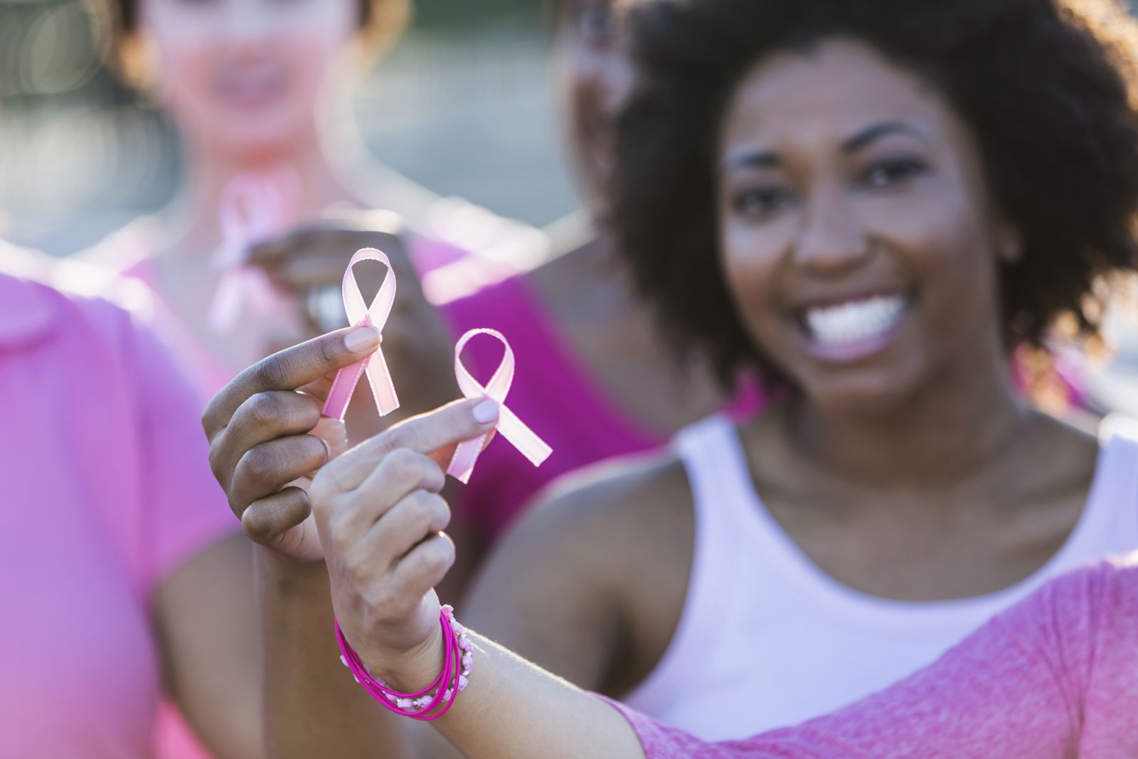 Cropped group of multi-ethnic teenage girls and young women holding breast cancer awareness ribbons in their hands, and wearing pink shirts. The focus is on the hand of the African American women who is smiling at the camera.