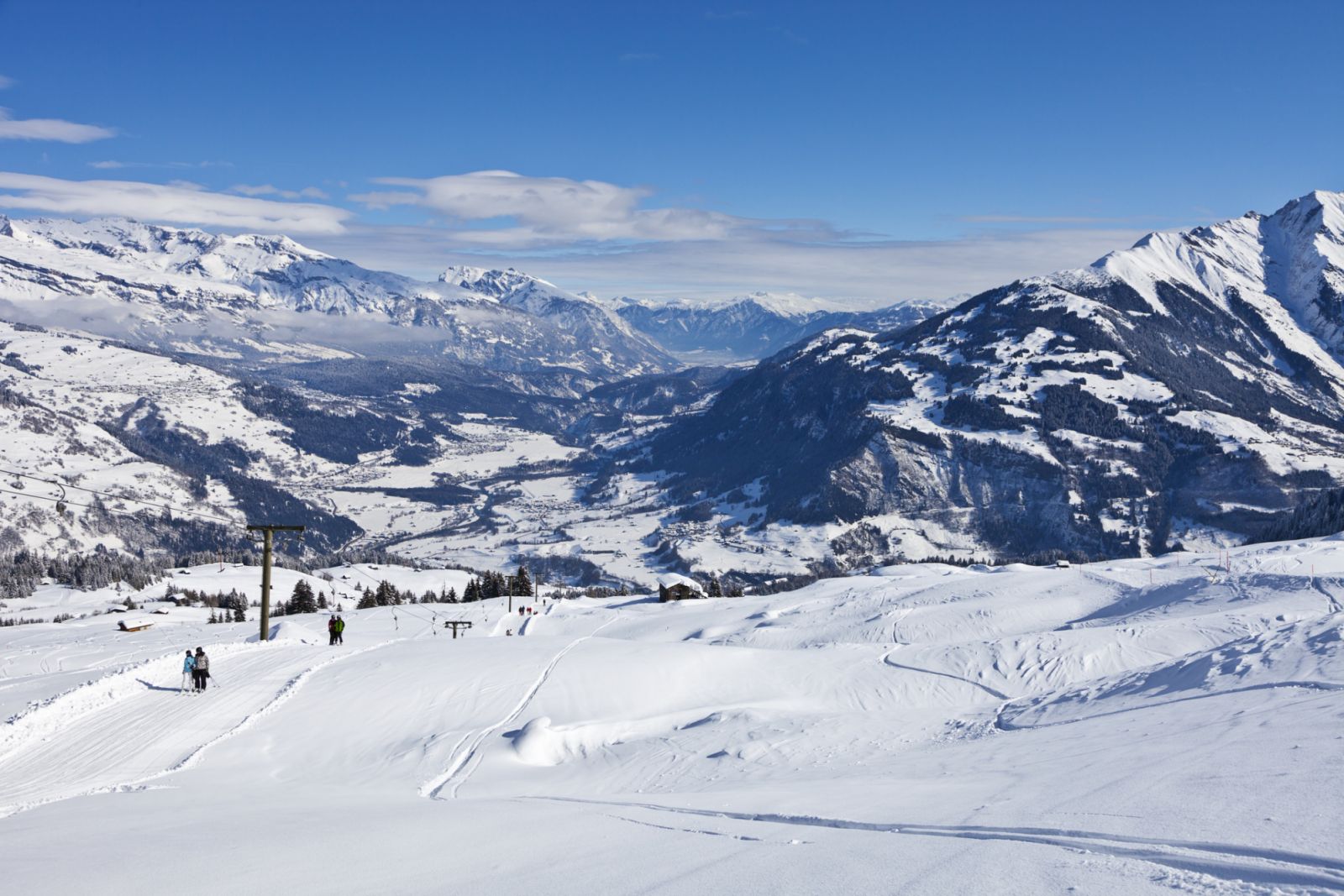 Unrecognizable skiers on a T-bar lift in the Obersaxen ski resort; Rhine valley with towns of Ruschein, Sagogn and Flims in background; in the far distance the city of Chur, capital of the canton of Graubuenden, can be seen