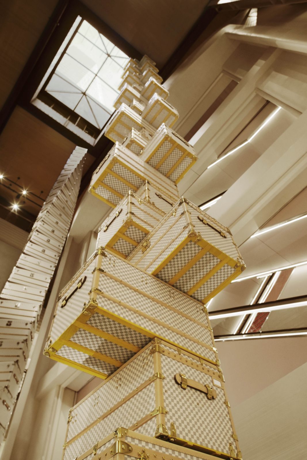 A detailed view of one of OMA’s trunk towers in white Damier pattern with gold hardware, demonstrating the architectural precision of the installation as it rises toward the skylight
