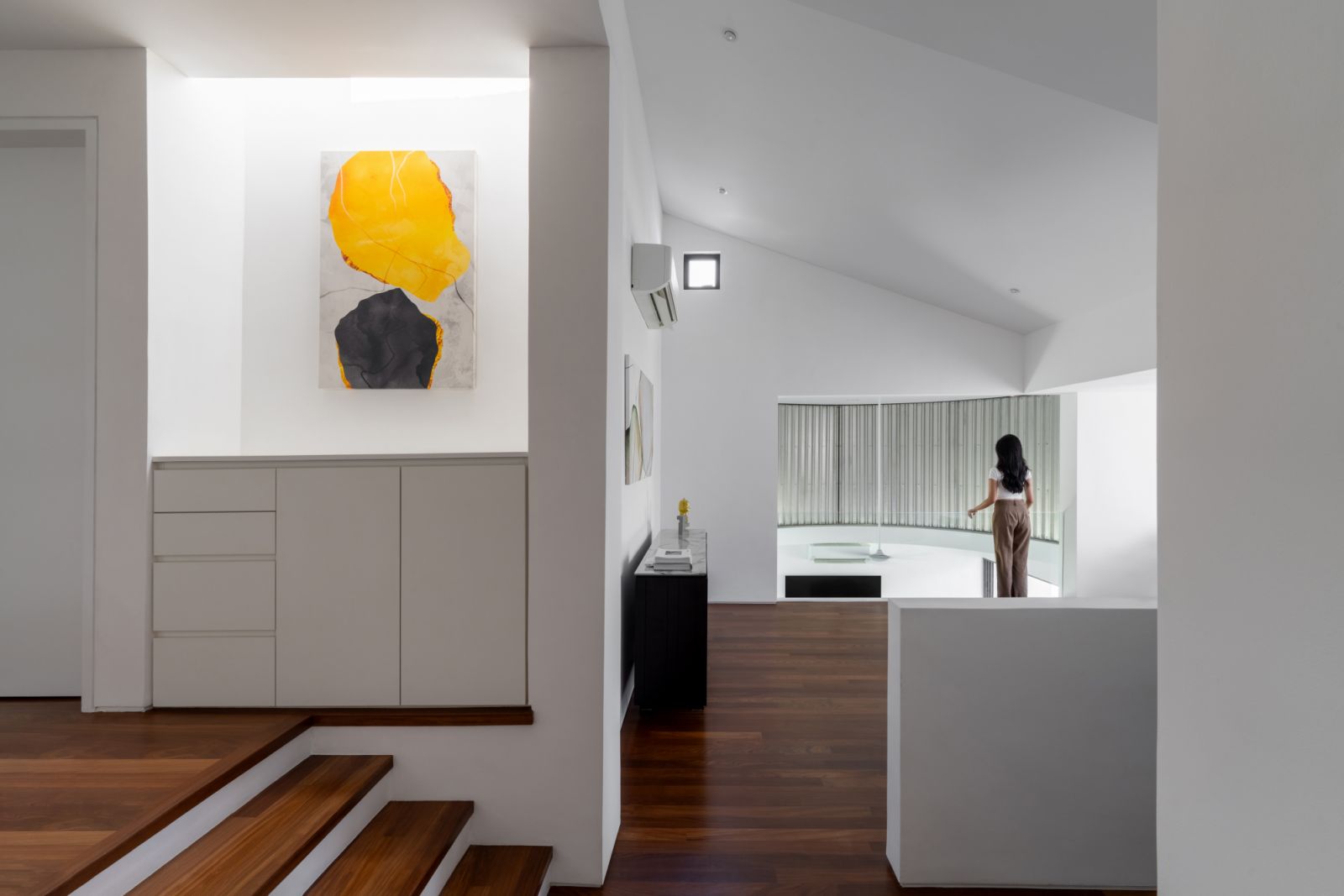The split-level interior showcases abstract artwork against white walls, while wooden steps and built-in storage maintain the home’s minimal aesthetic