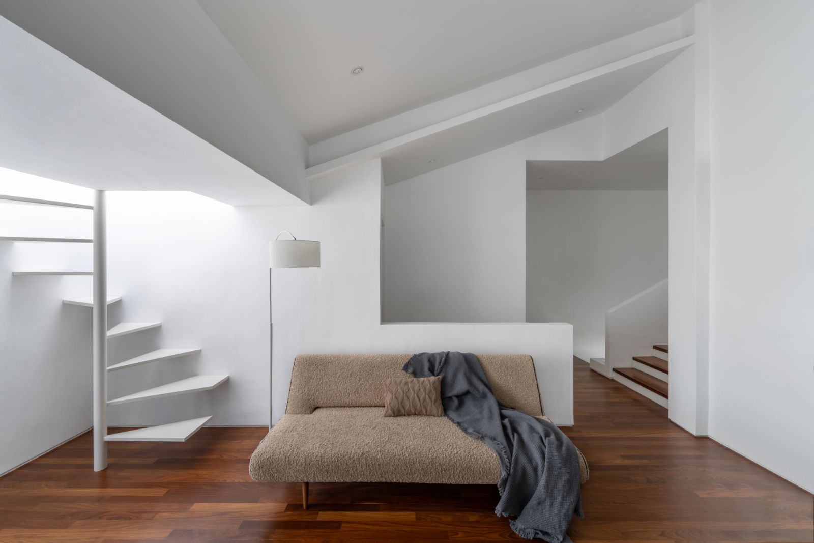 A secondary living space features a floating spiral staircase and bouclé daybed, creating a tranquil retreat. The minimalist design is enhanced by warm wood flooring and a strategically placed floor lamp