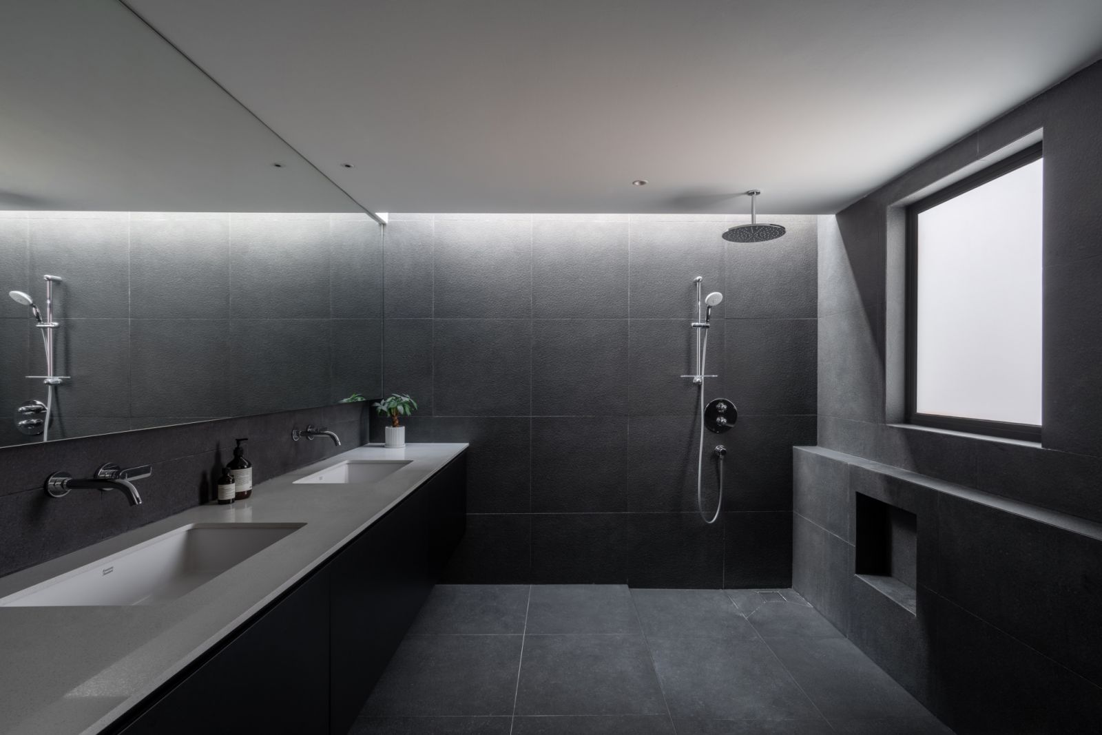 The minimalist bathroom features dark grey tiles and a floating double vanity, with natural light streaming in through a high window