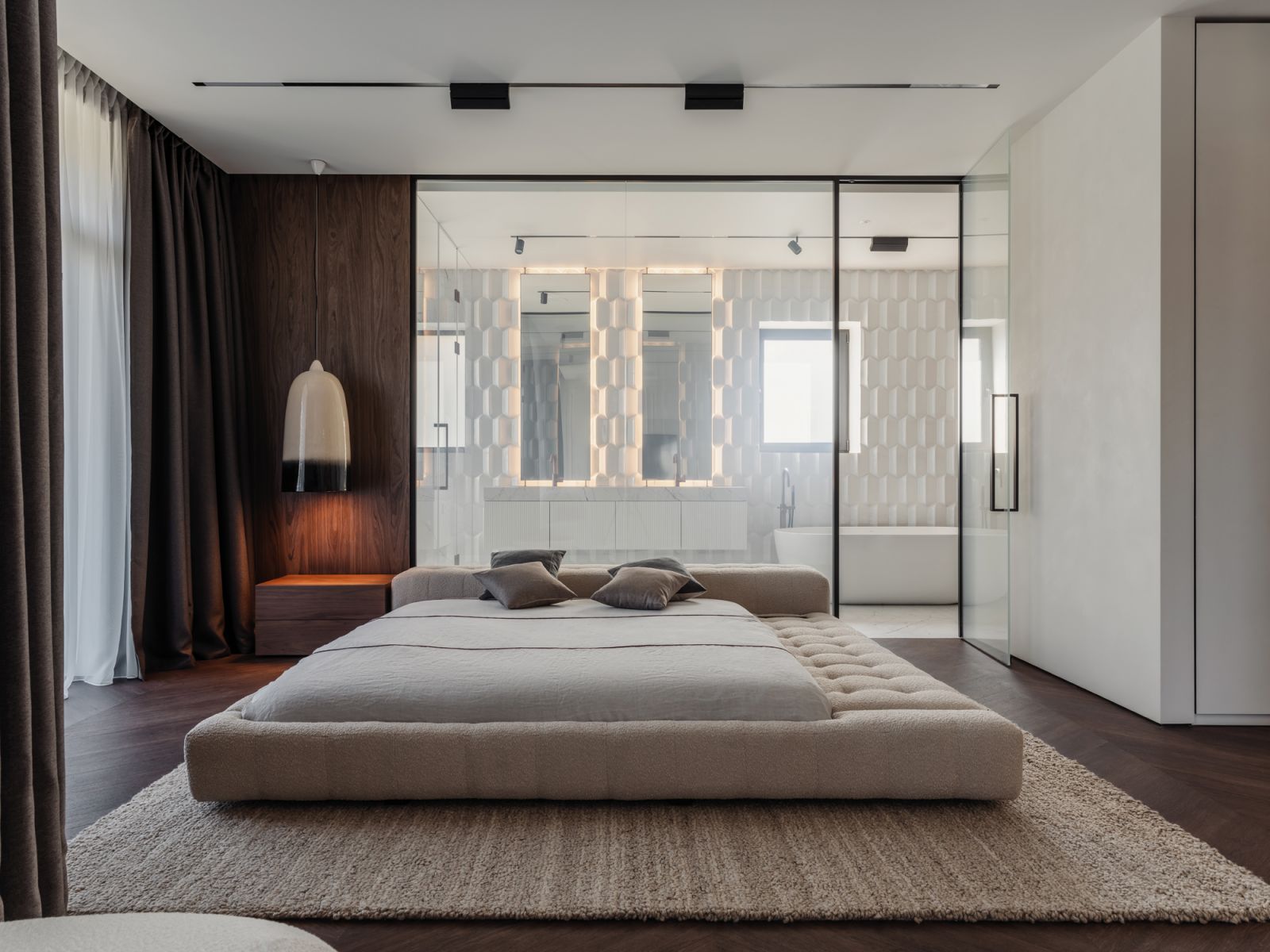 The master bedroom features a low-slung upholstered bed frame set against the bathroom’s glass partition