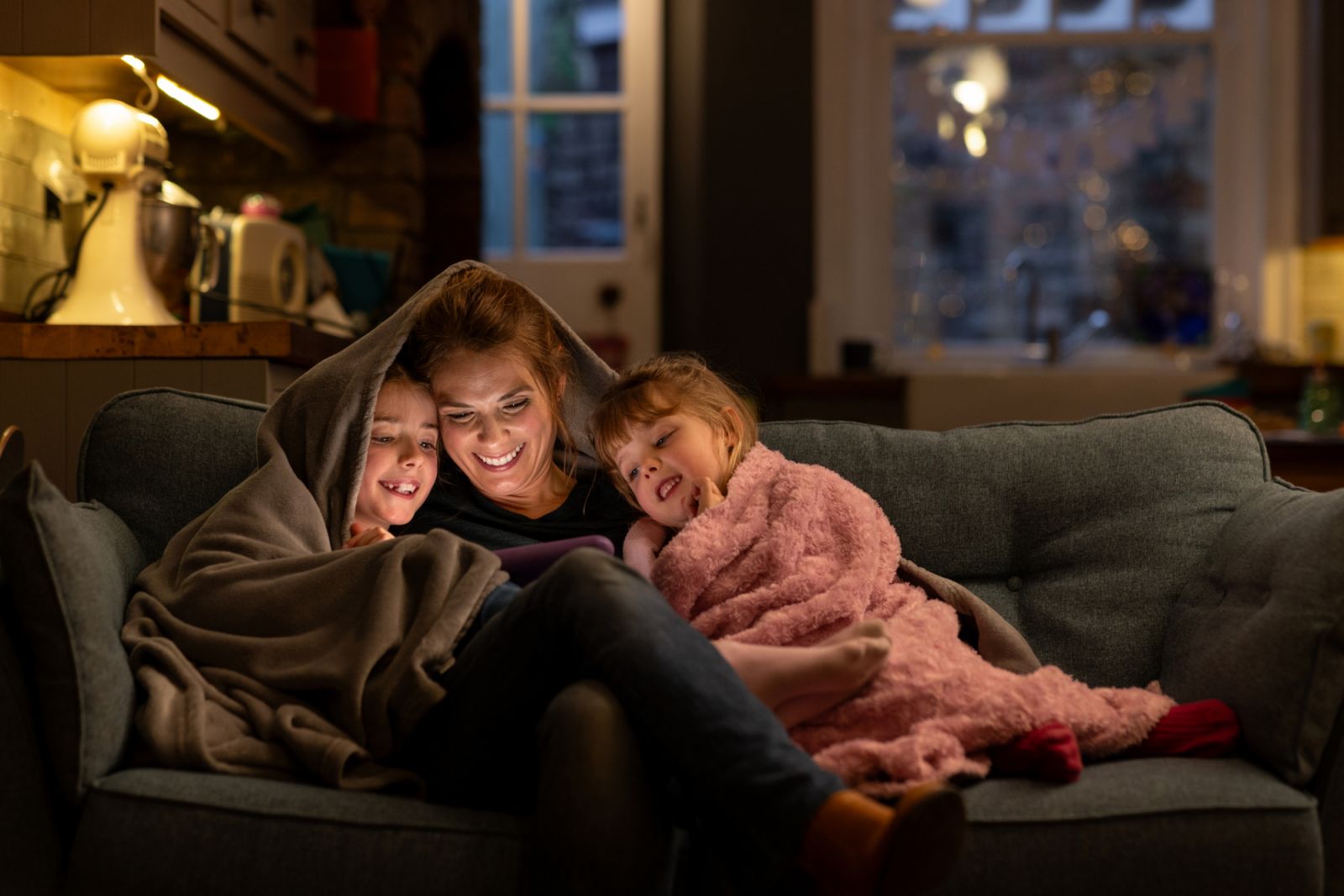 A shot of two young girls with their mother, sitting comfortably with a blanket over their heads on a sofa in the living room of their home in South Shields, North East England. They are all smiling, looking at a digital tablet which the mother is holding. In the background the kitchen is in partial view, and it is dusk outside.
Video similar to this scenario available.