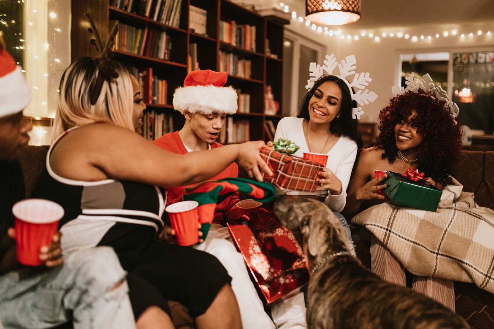 A group of young adult friends gather at a home for Christmas celebration over the holiday, dressed to fit the occasion with various Christmas accessories. They hand each other presents and open them cheerfully.