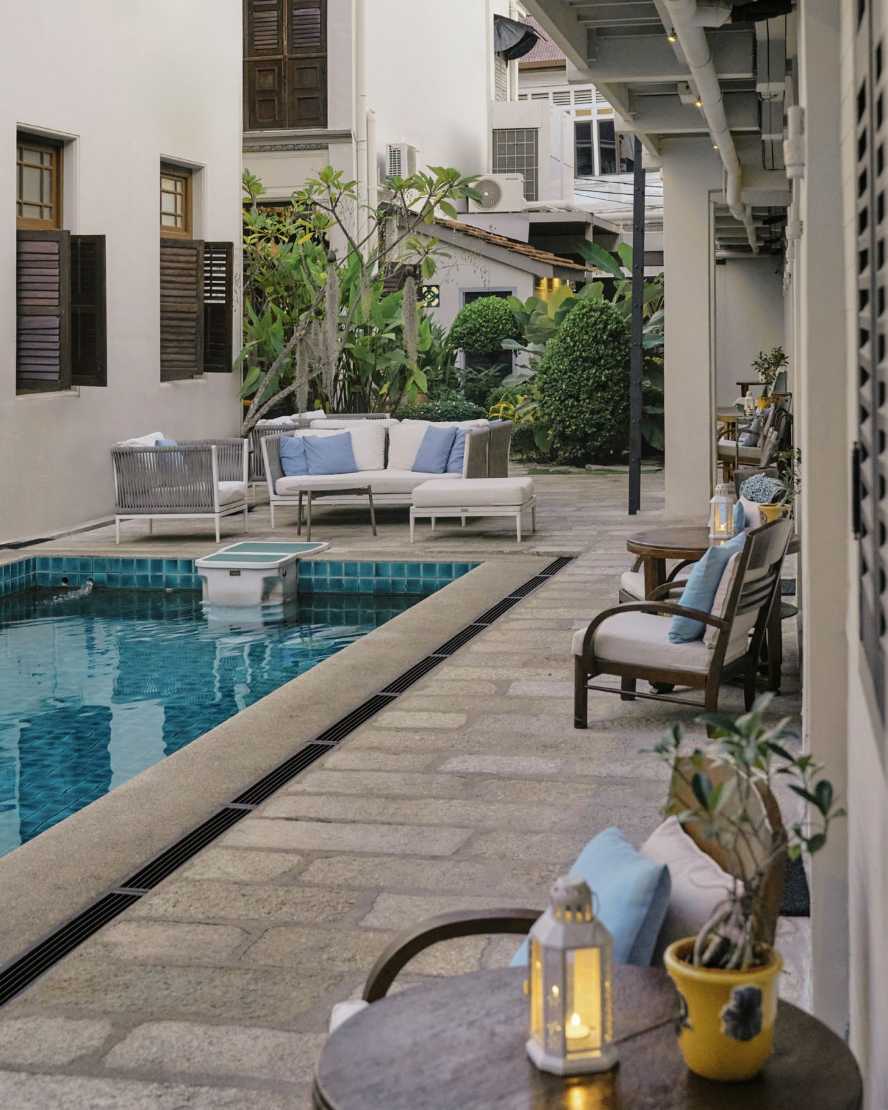 The tranquil courtyard pool area reflects Studio 11's modern interpretation of luxury, where contemporary furnishings in crisp whites and soft blues complement the historic shuttered windows and lush garden