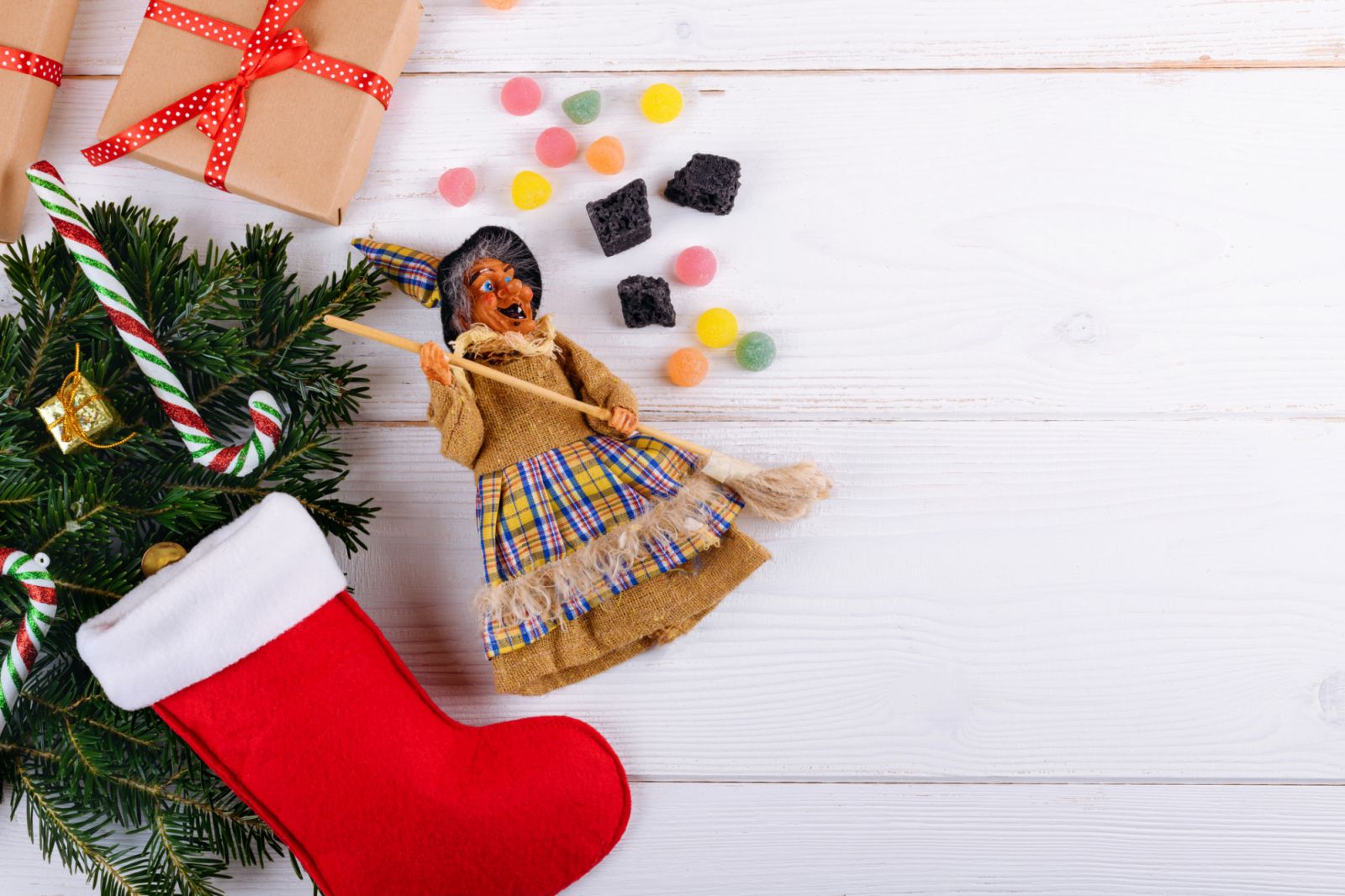 The witch Befana and red stocking with sweet coal and candy on white wooden background. Italian Epiphany day tradition, top view, flat lay, greeting card, copy space