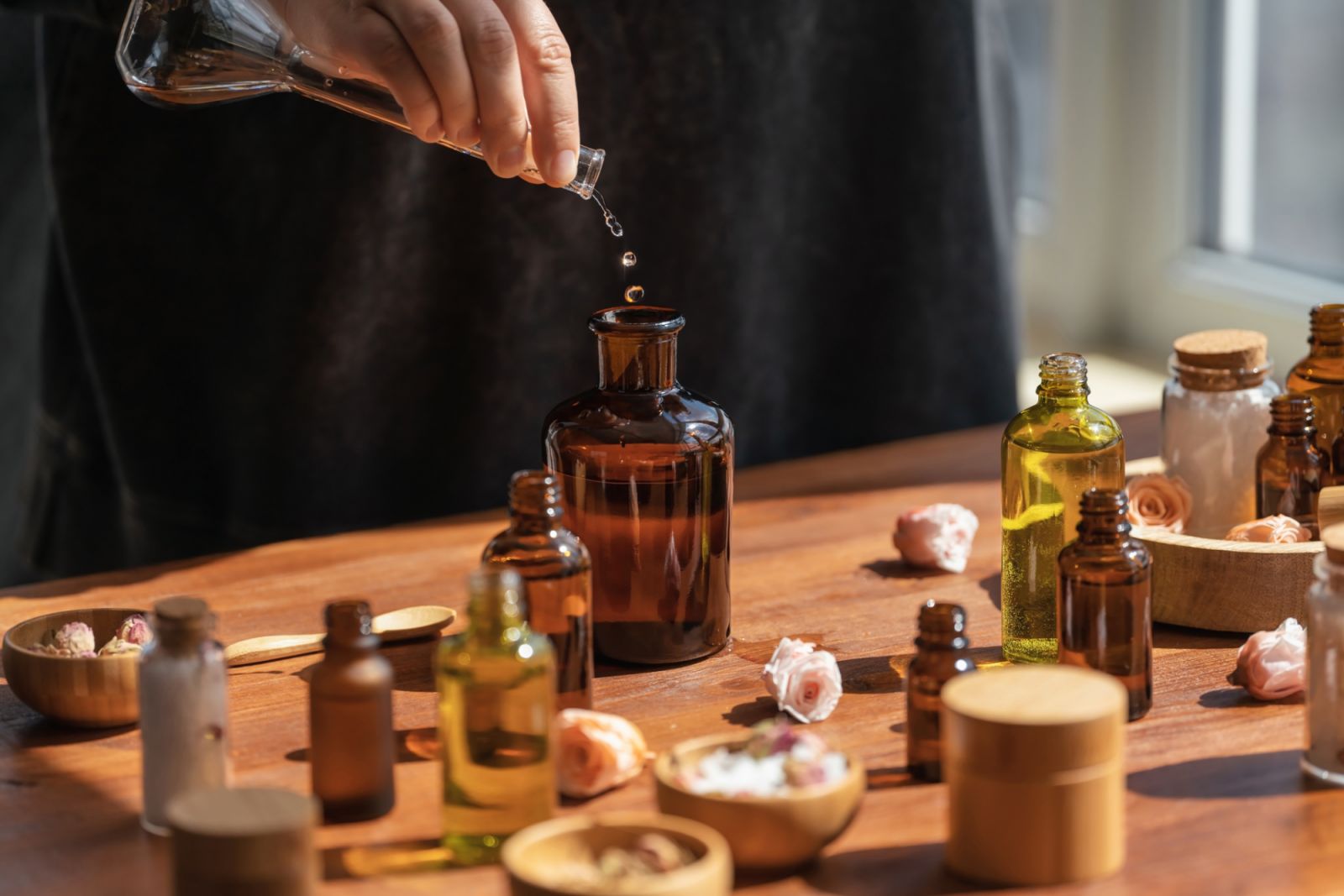 Woman preparing aromatic liquid for diffuser on wooden table. Homemade perfume from essential flower oil. Skincare products, natural cosmetic. DIY. Many bottles and jars with different types of oils, salt and essences. Alternative medicine concept.